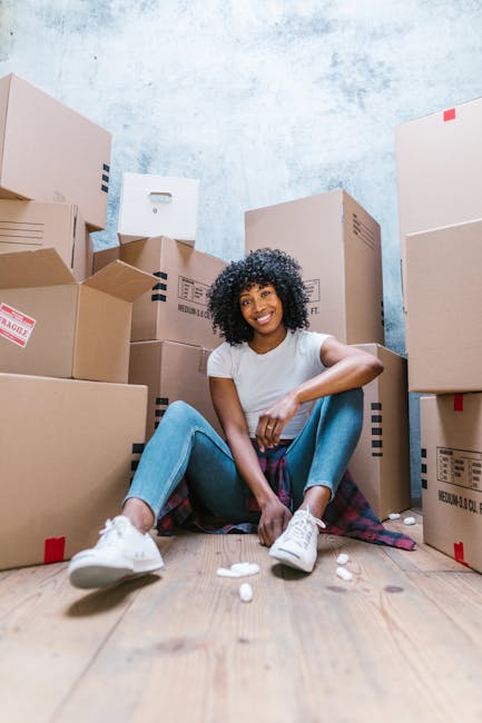 A young woman with curly black hair and a bright smile is sitting on a wooden floor inside a room, surrounded by numerous cardboard moving boxes of various sizes, some sealed with red or blue tape and labeled. She is wearing a white t-shirt, light blue jeans, and white sneakers, with a plaid shirt tied around her waist. The boxes are stacked in the background against a textured light blue wall, and the woman is casually leaning forward with her arms resting on her knees. Small packing materials, such as foam peanuts, are scattered on the floor around her. The scene depicts a home relocation or packing process, with the company Hackney Removals assisting with furniture transport, packing, and moving logistics, as suggested by the context of the image and the nearby boxes. The overall environment looks organized, indicating ongoing preparation for a house removal or storage service within the context of Dalston Removals and Storage by Dalston Junction.