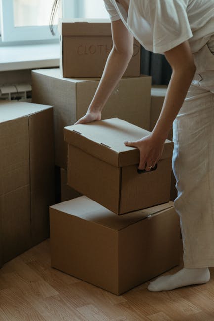 A person dressed in light-colored clothing is inside a room, stacking and organizing cardboard boxes used for packing during a home relocation. The individual is lifting a small box with plain brown surface and no visible labels, positioned on top of a larger box. Surrounding them are several other similarly plain cardboard boxes, some labeled with black marker, such as one marked 'Clothes' in the background. The boxes are arranged on a wooden floor near a window with natural daylight illuminating the scene. In the background, part of a table or furniture piece is visible. This image depicts the packing process as part of furniture transport and moving logistics, with the focus on careful handling and organization of household items during a house removal by Hackney Removals, in line with services offered at Dalston Junction.