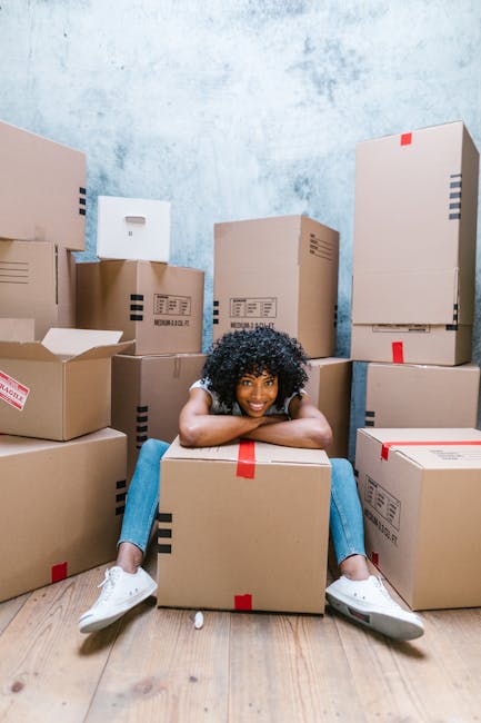 A young woman with curly black hair and a bright smile is sitting on a wooden floor inside a room, surrounded by numerous cardboard moving boxes of various sizes, some sealed with red or blue tape and labeled. She is wearing a white t-shirt, light blue jeans, and white sneakers, with a plaid shirt tied around her waist. The boxes are stacked in the background against a textured light blue wall, and the woman is casually leaning forward with her arms resting on her knees. Small packing materials, such as foam peanuts, are scattered on the floor around her. The scene depicts a home relocation or packing process, with the company Hackney Removals assisting with furniture transport, packing, and moving logistics, as suggested by the context of the image and the nearby boxes. The overall environment looks organized, indicating ongoing preparation for a house removal or storage service within the context of Dalston Removals and Storage by Dalston Junction.