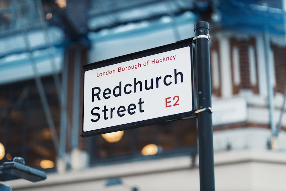 Close-up of a street sign mounted on a black pole displaying 'Redchurch Street' in large black text, with 'London Borough of Hackney' and 'E2' postcode in smaller red and black lettering; the sign is set against a blurred urban background with a building featuring large windows and a blue scaffold or metal framework, indicating an active city environment typical for house removals and home relocation logistics as provided by Hackney Removals; the scene is lit with natural daylight, highlighting the sign and surrounding structures, which are part of the moving process involving navigation through residential streets.