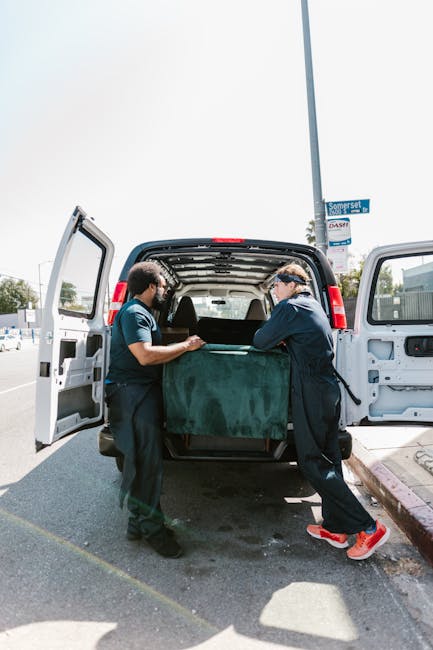 Two movers from Hackney Removals are engaged in a home relocation process, loading a large, dark green upholstered piece of furniture into the open rear of a white van parked on a street. The van's doors are wide open, revealing the metal interior and additional packing materials. Both movers are wearing dark uniforms with protective glasses; one is holding the furniture while the other guides it into the vehicle. The scene takes place outdoors on a paved street, with nearby signage and a clear sky overhead. Visible elements include the van, the furniture, protective gloves, and packing blankets or fabric wraps used for furniture protection. The environment includes a parking area adjacent to residential or commercial properties, emphasizing a professional furniture transport and packing process related to house removals and delivery services.