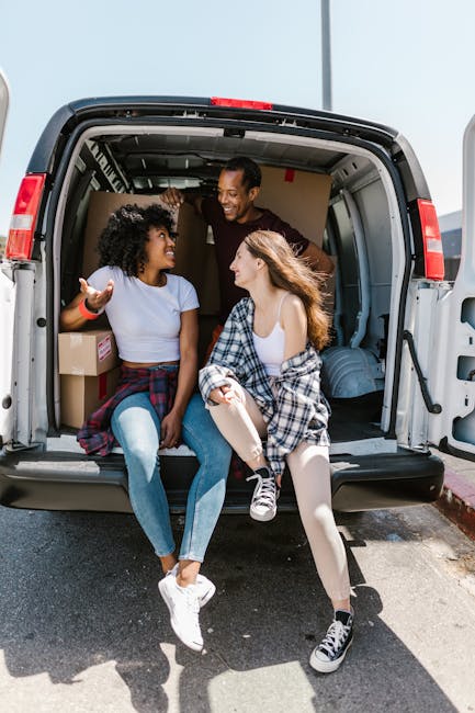 Two young women sitting at the edge of an open moving van during a house relocation, engaging in a friendly conversation. The woman on the left has curly dark hair, is wearing a white t-shirt and blue jeans, with a plaid shirt tied around her waist, and is gesturing with her right hand. The woman on the right has long, straight, light brown hair, is dressed in a white tank top and light-colored tights, with a plaid shirt draped over her shoulders, and is smiling while listening. Inside the van, visible cardboard boxes, some wrapped in plastic or packing paper, are stacked along with a large plastic container and soft furnishings. The van is parked on a paved street, and natural daylight illuminates the scene, suggesting the unloading process is part of a professional furniture transport or packing and moving service by Hackney Removals, specialising in house removals and studio moves. The environment reflects a typical urban residential area ready for furniture transport and home relocation.