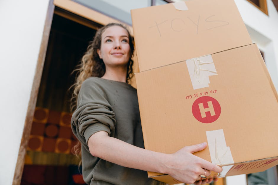 A young woman with long, curly hair and a neutral expression is inside a home, carrying two cardboard boxes stacked on top of each other. One box is labeled 'toys' in handwritten text, and both boxes are sealed with packing tape. The boxes are made of brown corrugated cardboard, and the woman is holding them at waist level with both hands. She is wearing a casual, long-sleeved gray top with rolled-up sleeves. The background shows an open doorway leading to a room with wooden paneling or artwork, indicating a home environment during a house removal or moving process, with visible details supporting packing and furniture transport activities. The image relates to home relocation, packing, loading, and furniture transport steps involved in professional removals services, like those offered by Hackney Removals.
