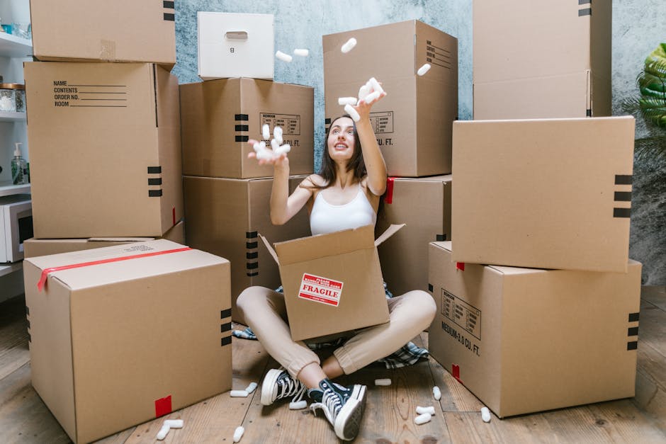 A young woman with long dark hair, sitting on the floor of a room surrounded by numerous assorted cardboard boxes used for packing during a home relocation. She is smiling and looking up as she throws small white packing peanuts into the air. The boxes vary in size, some sealed with red and black tape, and are stacked against the light blue and grey textured wall. The woman is dressed casually in a white sleeveless top, beige pants, and black-and-white sneakers. Inside the open box on her lap, a red label marked 'FRAGILE' is visible, indicating careful handling of delicate items. The room has a wooden flooring, and a small kitchenette with utensils and a potted plant can be seen in the background. This scene illustrates the packing and moving process, characteristic of furniture transport and home relocation efforts, with an emphasis on the importance of organized packing for house removals as provided by Hackney Removals.