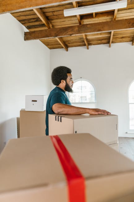 A man with short curly hair and a beard, wearing a teal shirt, stands inside a spacious room with white walls and a high wooden ceiling supported by exposed beams. He is positioned behind several large cardboard boxes, one of which has red and black packing tape, indicating they are packed for a home relocation. The boxes are stacked and arranged on the floor, with some boxes placed on a trolley or moving cart. The man is resting his hands on the top of one box and appears to be preparing for or engaged in the loading process. The room features three arched windows allowing natural daylight to illuminate the space, with a view of neighbouring buildings visible through the windows. This scene exemplifies packing and furniture transport during a professional house removal carried out by Hackney Removals, reflecting careful handling of belongings in preparation for or during a move within the Hackney area.