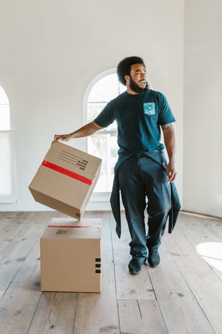 A young woman with long dark hair, sitting on the floor of a room surrounded by numerous assorted cardboard boxes used for packing during a home relocation. She is smiling and looking up as she throws small white packing peanuts into the air. The boxes vary in size, some sealed with red and black tape, and are stacked against the light blue and grey textured wall. The woman is dressed casually in a white sleeveless top, beige pants, and black-and-white sneakers. Inside the open box on her lap, a red label marked 'FRAGILE' is visible, indicating careful handling of delicate items. The room has a wooden flooring, and a small kitchenette with utensils and a potted plant can be seen in the background. This scene illustrates the packing and moving process, characteristic of furniture transport and home relocation efforts, with an emphasis on the importance of organized packing for house removals as provided by Hackney Removals.