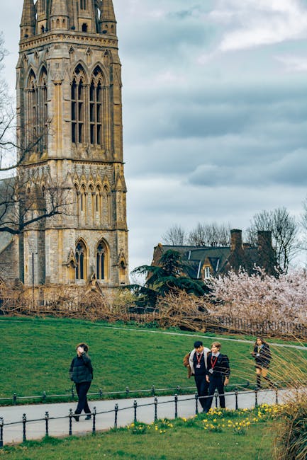 A group of four people dressed in casual and business attire walking along a paved pathway in front of a historic stone church or cathedral with tall, detailed gothic-style architecture, large stained glass windows, and multiple towers. The scene is set outdoors on a cloudy day, with surrounding greenery including a sloping grassy area, flowering bushes, and trees in the background. The image captures the urban environment of Stoke Newington on Church Street and exemplifies a typical day with pedestrians in a local community area, aligning with themes of home relocation or moving logistics. The setting demonstrates an environment where furniture transport and packing during house removals may occur, reflecting services offered by Hackney Removals, including loading or unloading activities in an outdoor or street-based setting.