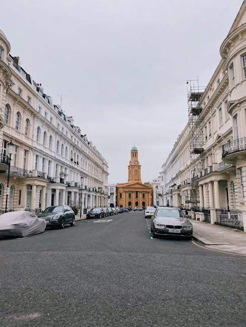 Photograph taken during daytime showing a quiet residential street in Stoke Newington, with a view towards a historic building with a clock tower and a dome at the end of the road. The street is lined with white Victorian-style terraced houses featuring wrought iron balconies and large windows. Several parked cars, including a covered vehicle, are on the paved road, and some vehicles are positioned near the curb. Inside the street, a Hackney Removals team is engaged in a home relocation process, with visible packing materials such as cardboard boxes, bubble wrap, and furniture blankets on a trolley near the entrance of a property. The team appears to be preparing to load or unload furniture and packed items into a moving van stationed nearby, with some items being carried or positioned close to the house doorway. This scene exemplifies furniture transport and packing during house removals in Stoke Newington, combining the urban environment with professional moving logistics.