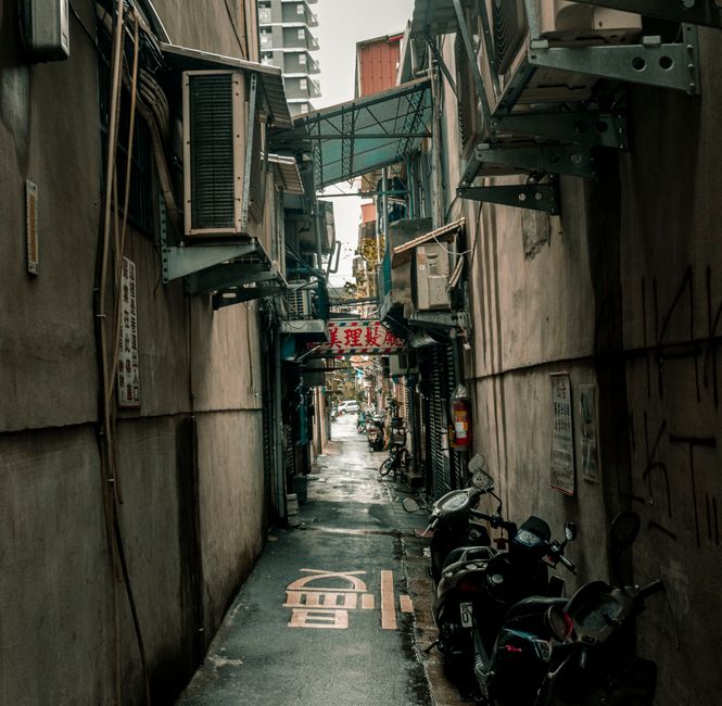 A narrow alleyway between two brick buildings with a paved pathway made of rectangular stone slabs. The walls of the buildings are constructed from red and dark brown bricks, with some pipes and elect
